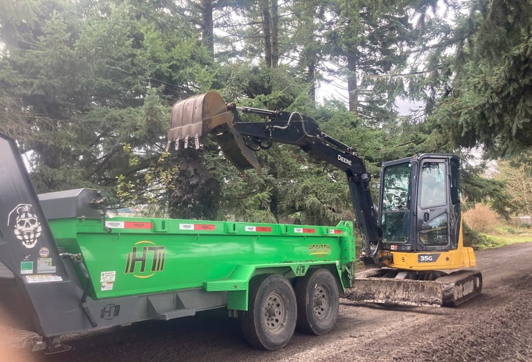 Mini excavator loading wood debris into green dump trailer on forest road
