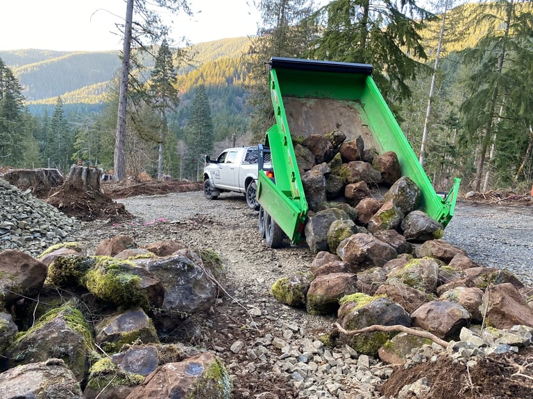 Bright green tipper truck dumping rocks and boulders in a forest setting with mountains and evergreen trees in the background
